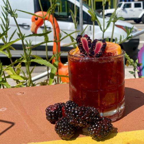 A refreshing cocktail garnished with blackberries and an orange slice, with a flamingo decoration in the background.