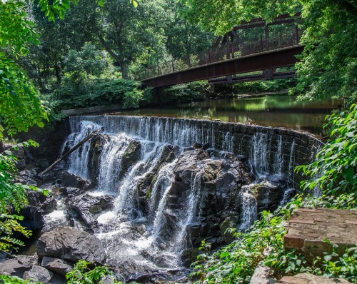 A serene waterfall cascades over rocks, surrounded by lush greenery, with a bridge in the background.