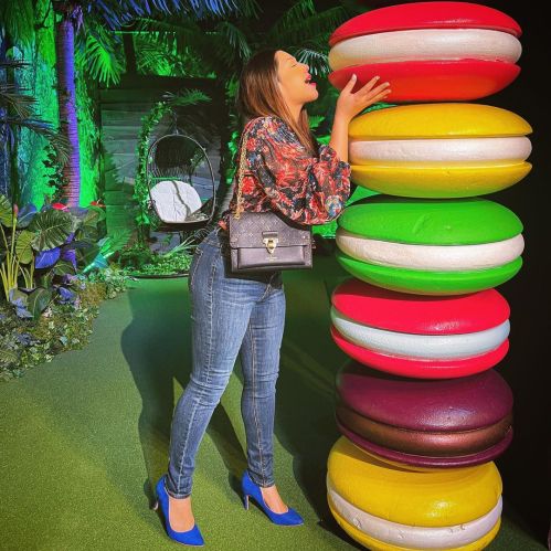 A woman in a floral top and jeans playfully leans towards a colorful stack of oversized macarons in a vibrant setting.