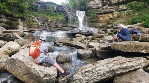 Two people sit on rocks by a waterfall, surrounded by greenery and flowing water in a serene outdoor setting.
