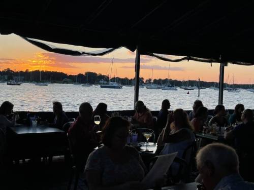 People dining at a waterfront restaurant during sunset, with boats moored in the background.