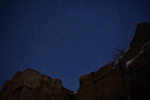 A starry night sky viewed between rocky cliffs, with a few scattered clouds and snow on the ground.