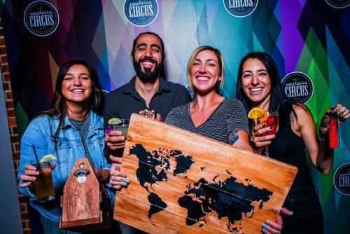 Four friends smile and pose with drinks and a wooden world map against a colorful backdrop.