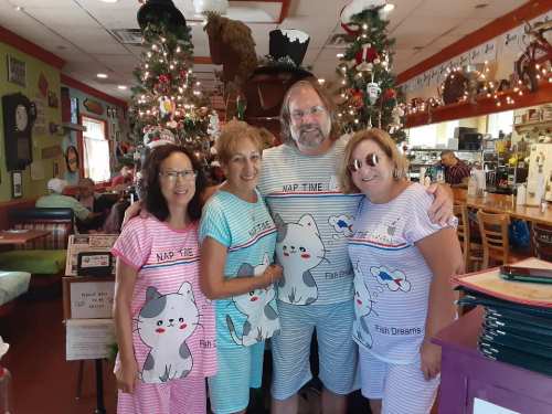 Four people in matching cat-themed pajamas pose together in a festive restaurant decorated with Christmas trees.
