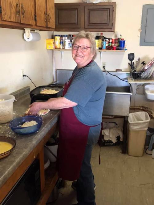 A smiling woman in an apron prepares food in a cozy kitchen, surrounded by bowls and cooking utensils.