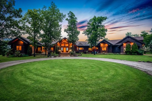 A beautiful log cabin home surrounded by trees, with warm lights glowing at dusk and a circular lawn in front.