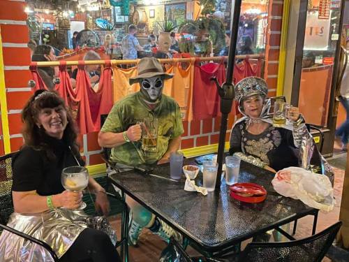 Three people sitting at a table in a colorful restaurant, enjoying drinks and wearing festive outfits.