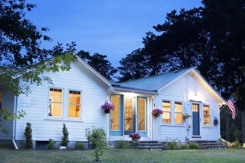 A charming white house at dusk, with warm lights glowing from the windows and flowers adorning the entrance.