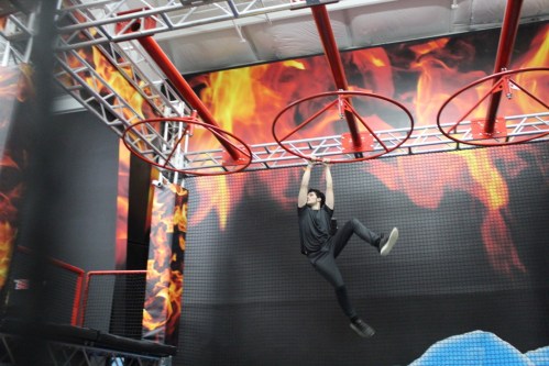 A person hangs from a red obstacle course ring in a gym with a fiery backdrop.