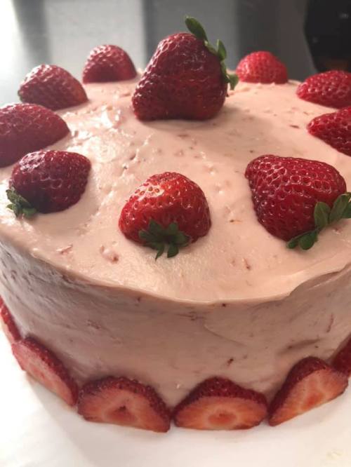 A round cake topped with fresh strawberries and a smooth pink frosting, decorated with sliced strawberries around the base.