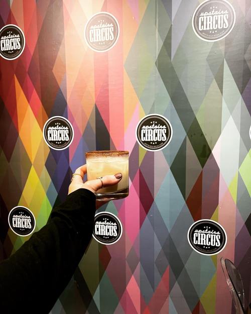 A hand holding a drink in front of a colorful, diamond-patterned wall with "Majestic Circus" logos.