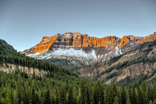 A stunning mountain range with snow-capped peaks and vibrant orange hues, surrounded by lush green forests.