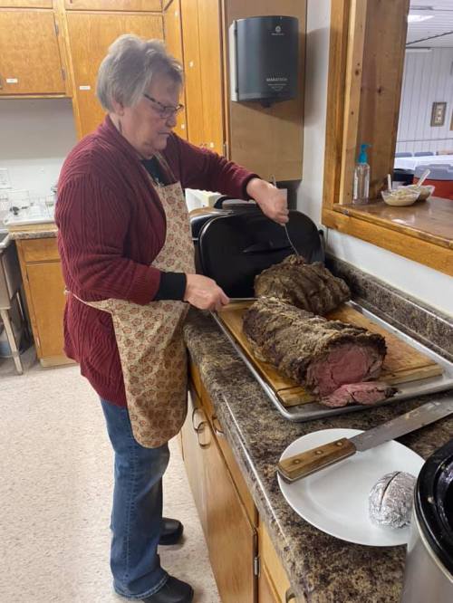 A woman in an apron slices a large roast on a kitchen counter.
