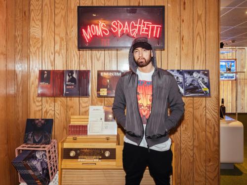 A person stands in front of a neon sign for "Mom's Spaghetti," surrounded by music albums and a vintage radio.