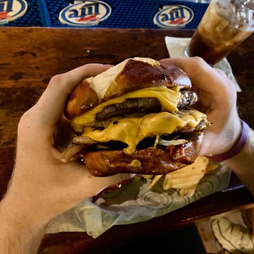 A person holds a large, cheesy burger with two patties on a pretzel bun, with a drink in the background.