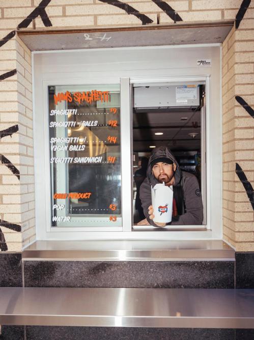 A man in a hoodie holds a takeout container at a restaurant window with a menu for "Mom's Spaghetti."