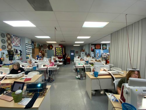 A spacious sewing room with several people working on sewing machines, surrounded by colorful quilts and fabric.