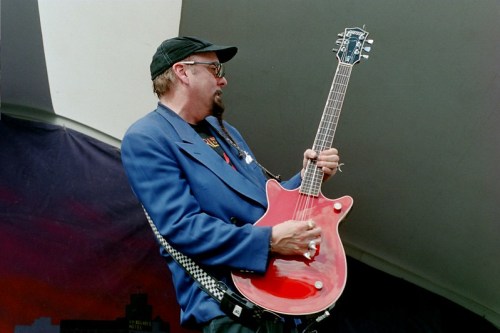 A musician in a blue jacket and cap plays a red guitar on stage, passionately performing.