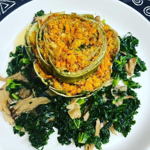 Stuffed artichoke on a bed of sautéed kale and mushrooms, garnished with herbs and spices.