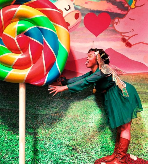 A person in a green dress and fairy wings playfully reaches for a large, colorful lollipop against a whimsical backdrop.