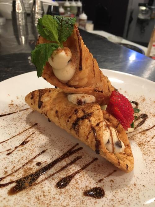 Two crispy cannoli filled with cream, garnished with a mint leaf and a strawberry, drizzled with chocolate sauce.