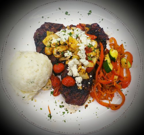 A plated meal featuring grilled steak topped with sautéed vegetables, feta cheese, and a side of mashed potatoes and salad.