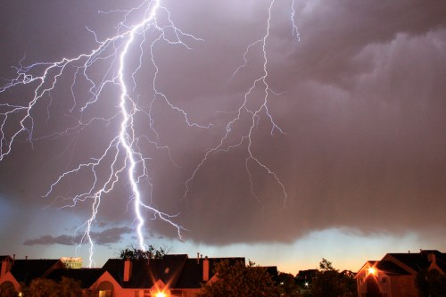 A dramatic lightning storm illuminates a dark sky above rooftops, with streaks of lightning striking down.