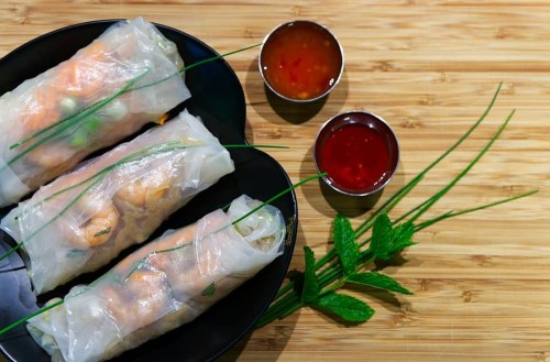 Three fresh spring rolls on a black plate, accompanied by dipping sauces and garnished with mint and chives.