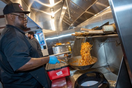A chef in a black hat stirs noodles in a commercial kitchen, with steam rising from pots in the background.