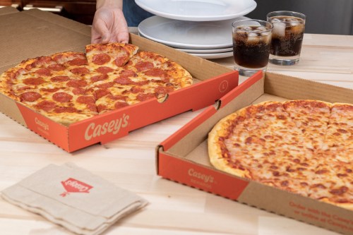 A person serves pepperoni pizza from two boxes, with drinks and plates in the background on a wooden table.