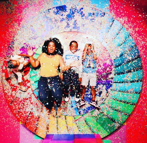 A group of children and an adult joyfully jumping in a colorful, confetti-filled setting.