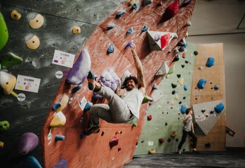 A climber hangs from a colorful indoor bouldering wall, while another person climbs in the background.