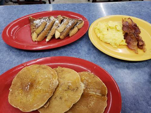 A colorful breakfast spread featuring French toast, scrambled eggs, bacon, and pancakes with syrup on red and yellow plates.