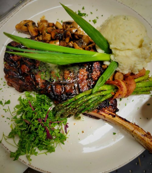 A grilled steak with mushrooms, mashed potatoes, asparagus, and garnishes on a white plate.