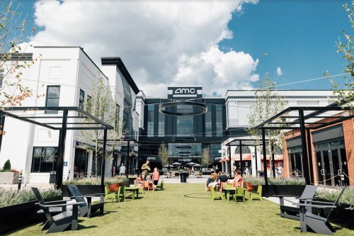 A vibrant outdoor area with green grass, seating, and people enjoying a sunny day near an AMC theater.