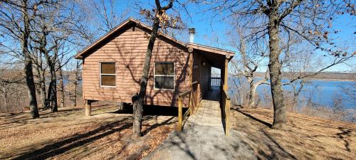 A wooden cabin on stilts surrounded by trees, overlooking a calm lake under a clear blue sky.