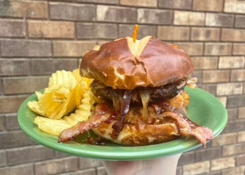 A pretzel bun burger topped with bacon and onions, served with a side of potato chips on a green plate.