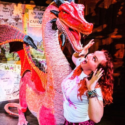 A woman with red hair poses playfully next to a large, colorful dragon sculpture in a whimsical setting.