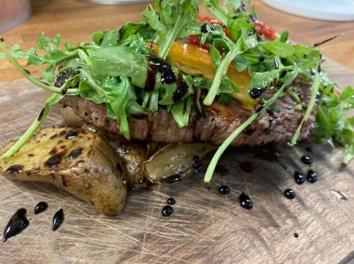A juicy steak topped with fresh greens and colorful peppers, served on a wooden board with roasted potatoes.