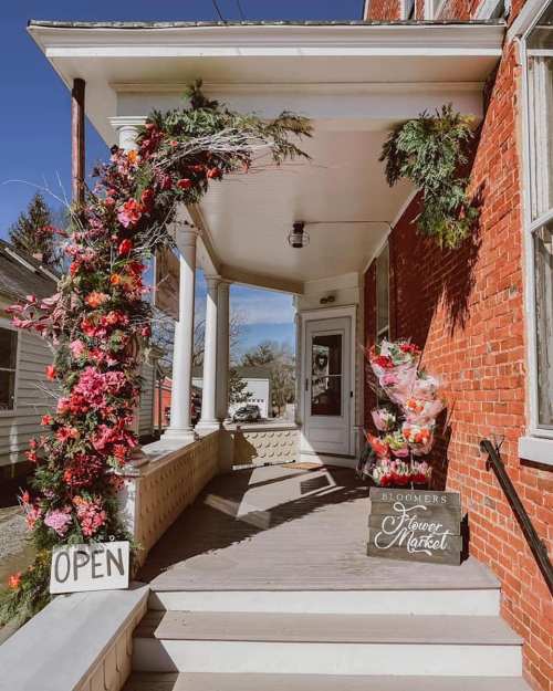 A charming brick entrance adorned with flowers and greenery, featuring an "Open" sign and a flower market display.