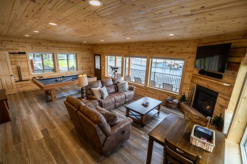Cozy wooden living room with a pool table, fireplace, and large windows overlooking a scenic view.