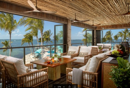 Cozy outdoor seating area with a view of the ocean, palm trees, and a pool, featuring natural decor and bright sunlight.