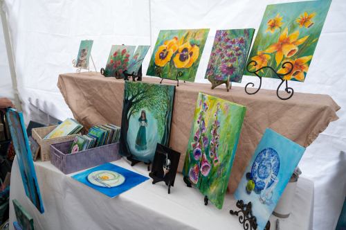 A display of colorful paintings on easels, featuring flowers and abstract designs, set up on a table under a tent.