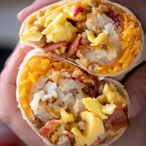 A close-up of a hand holding a cut burrito filled with eggs, cheese, bacon, and potatoes.