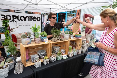 A vendor hands a bag to a customer at a market stall displaying various potted plants and planters.