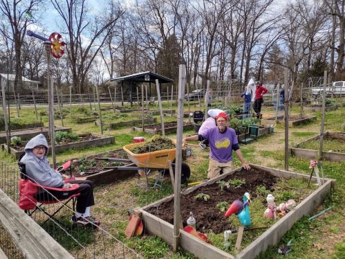 A community garden with people planting and tending to crops, while one person relaxes in a chair nearby.