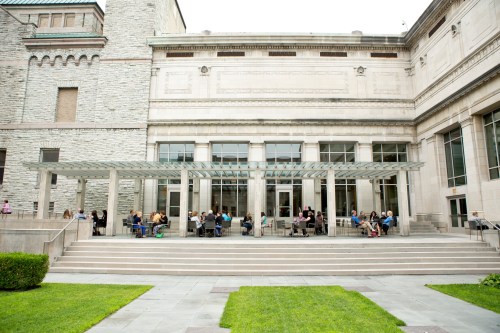 A modern building with a glass canopy, featuring people seated at tables outside on a patio surrounded by greenery.