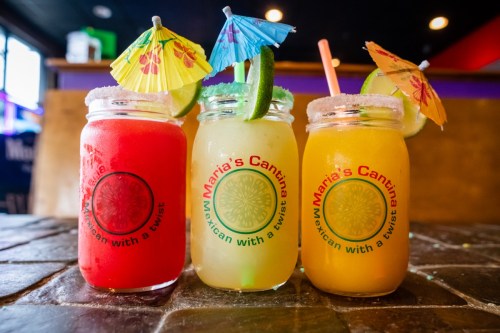 Three colorful cocktails in jars, each with a small umbrella and garnished with lime, on a rustic table.