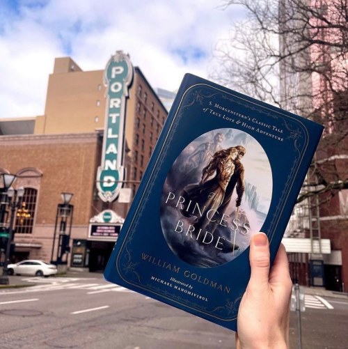 A hand holds "The Princess Bride" book in front of a vintage Portland sign and a city street.