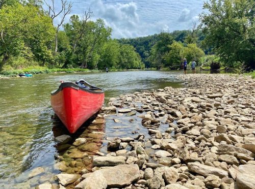 A red canoe rests on rocky shore by a river, with trees and people fishing in the background under a cloudy sky.